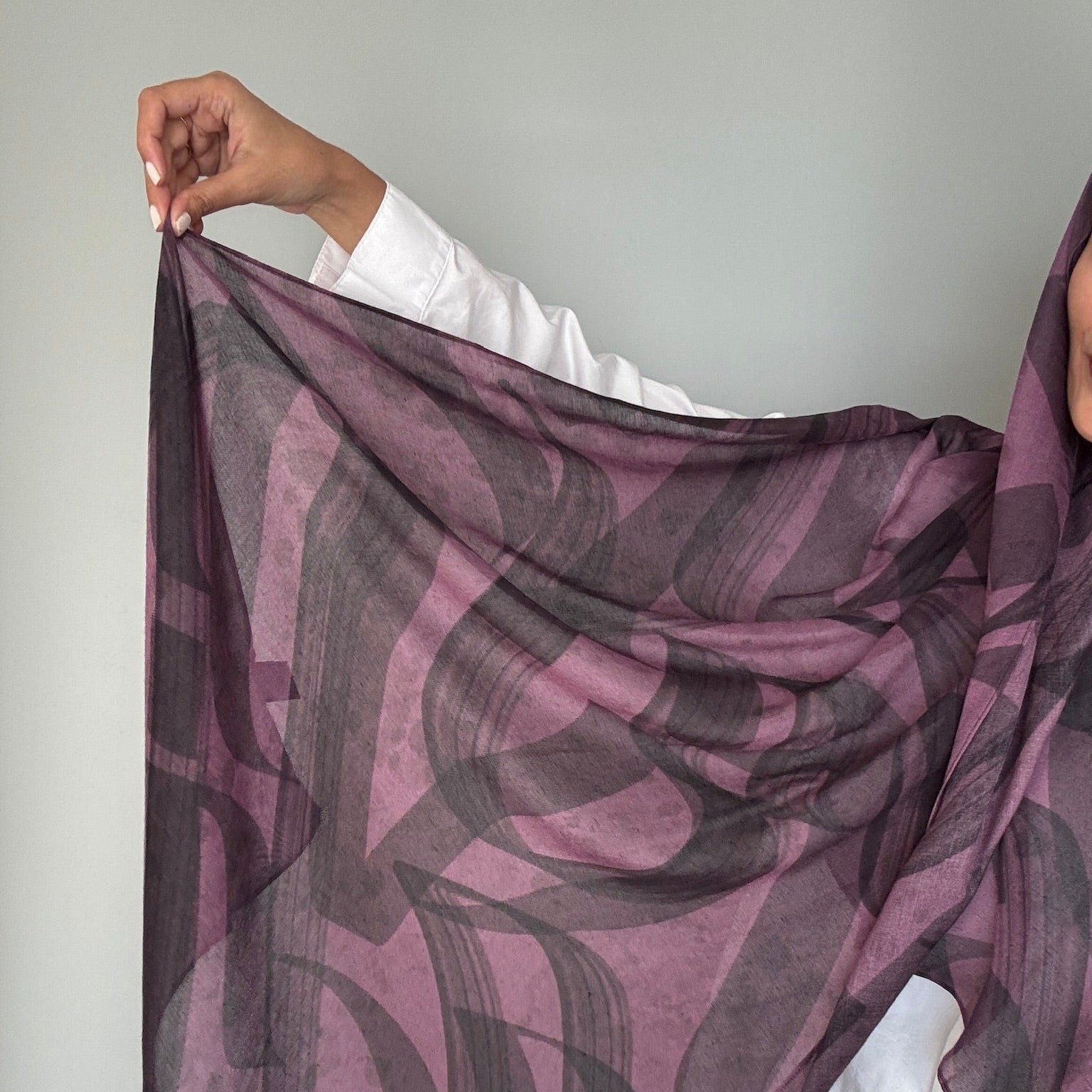 A person holds up a hijab with an abstract brushstroke pattern in dusty purple and dark gray, worn over a white shirt against a plain light gray wall.