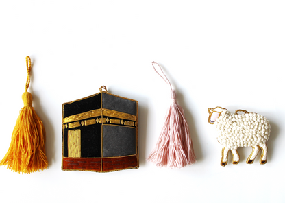 A collection of various handmade ornaments including representations of religious and cultural symbols such as the Kabaah, a mosque, a Quran, and other significant items for Muslims, along with tassels and a gold display cord.